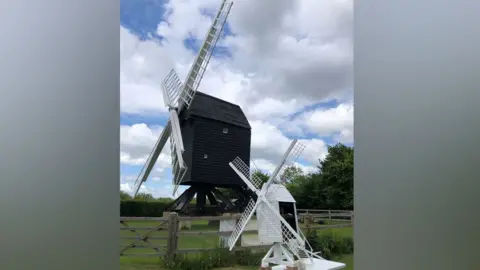 Cambridge Past, Present and Future Two wooden mills. The one in the background if a full size black-bodied wooden mill, sideways on with its white sails open in an X shape. The one in front is a white miniature replica, resting on a table. The mills are in fields under white clouds and blue sky.