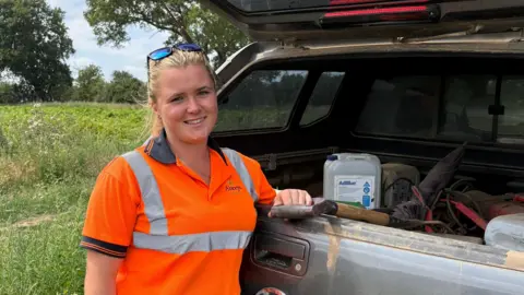 Ben Schofield/BBC Bonnie Oakley looking directly down the camera. She is standing at the back of a vehicle, whose tailgate is open. She is holding the handle of a garden fork that is lying inside the boot, alongside other equipment. She is wearing a bright orange, high visibility T-shirt. She is smiling and has a pair of sunglasses on her head. Her dark blonde hair is pulled back behind her head. She and the vehicle are in a field, with trees and a partly cloudy sky in the background. 