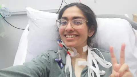 Submitted Mehreen Ahmed in a hospital bed with tubes around her neck following her heart transplant. She is wearing glasses and a green gown and is smiling into the camera with her fingers raised in a V for victory sign.