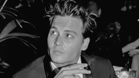Richard Blanshard A black and white image of Johnny Depp holding a cigarette. He is wearing a tuxedo and has tousled hair. 