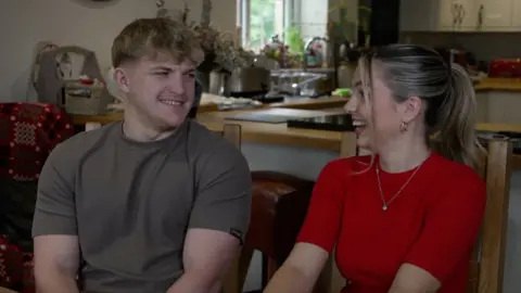 Race Across the World contestants Fin and Sioned smile at eachother inside a kitchen. 