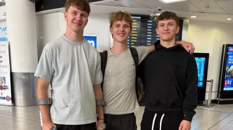 supplied Three young men pose together in an airport lounge. The man on the left is using two crutches to support himself.