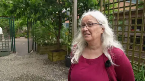 Marie-Louisa Niehus wearing a dark pink top and cardigan. She is shoulder length white hair and glasses. She is stood in front of a wooden fence and a tree. 