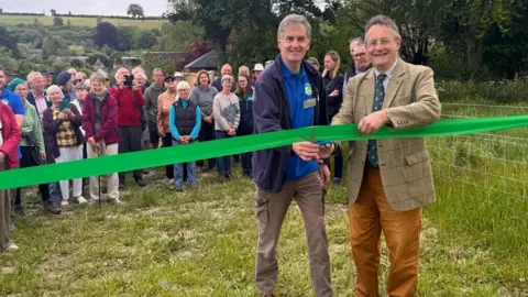 Wiltshire Council Two men, one in a brown tweed suit, another in a blue polo top and navy hoodie, holding a pair of scissors and cutting a green ribbon. There is a crowd of people behind them smiling and taking photgraphs. They are all standing outside in a field.