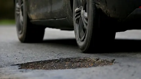 The wheels of a car, viewed from a low angle, travel past a pothole in the road. The car is black, and has mud splattered on its sides.