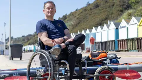 Will sat in his wheelchair with a paddleboard and equipment behind him on the beach