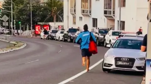 A lifeguard runs down a road barefoot carrying a red defribrillator bag next to parked cars on a road with trees and the sea in the background.