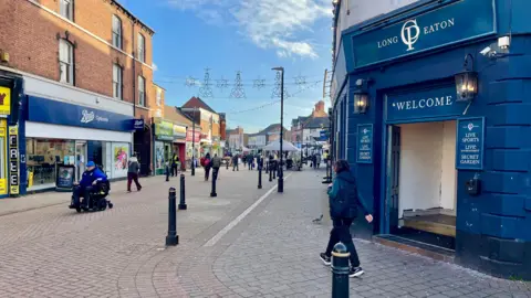 A town centre with a pedestrianised area between a pub called "Long Eaton" and a Boots chemist. There are Christmas lights hanging above the area, black bollards lining the pavement and people walking including a woman in a navy coat and a man in a blue hat driving an electric wheelchair. More shops and market stalls can be seen in the distance.