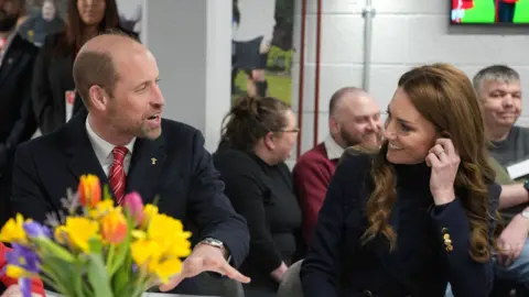 Prince William and Princess Catherine engaged successful  speech  astatine  a table. He has his manus  retired  and she is holding her near  ear. A vase of flowers is disposable   connected  the array  successful  beforehand   of them and radical   are talking and smiling successful  the background