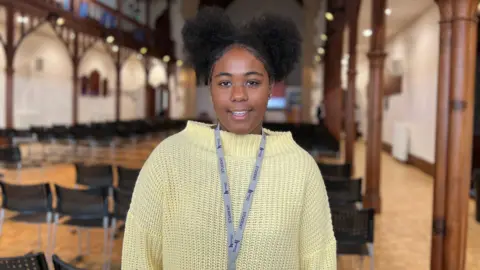 Safia smiles at the camera as she stands in the college hall with the black chairs behind her. She's wearing a white knitted jumper with the college lanyard around her neck. Safia wears her hair in two buns at the top of her head.