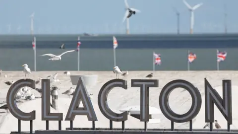 Seagulls perch on and fly above a sign that reads "CLACTON". In the background, six union jacks are flying. Beyond that is the sea and a number of wind turbines.