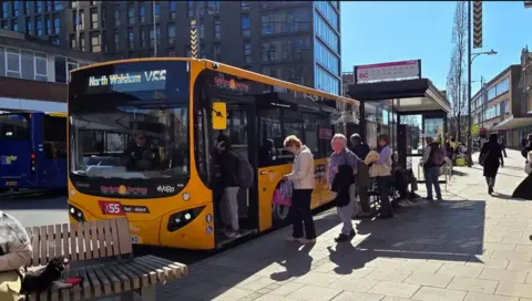 Passengers at a bus stop in Norwich waiting to get on a yellow bus travelling to North Walsham. It is a sunny day.