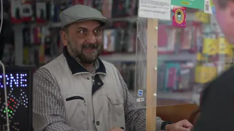 Hussein wears a grey baker boy cap, beige workman gilet and a black and beige check skirt. He stands inside his mobile shop.
