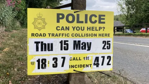 A photo showing a yellow police board attached to a fence post next to a road. There is black writing on the board appealing for witnesses to an accident 