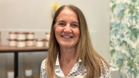 A woman with long light coloured hair, wearing a cream apron over a patterned blouse. She is smiling at the camera. Behind her are candles on a shelf