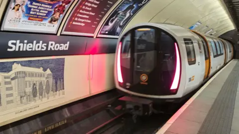 A white and orange subway train pulling away from Shields Road station.
