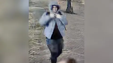 The image shows a blurry CCTV image of a man in a grey jacket and black bottoms. He is walking and the trunk of a tree can be seen in the background.