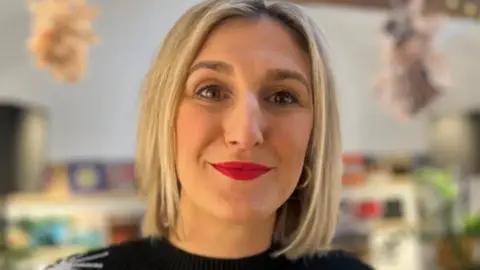 A close-up of Fliss Loxley. She has shoulder length light brown hair and is smiling. She is wearing a black jumper.