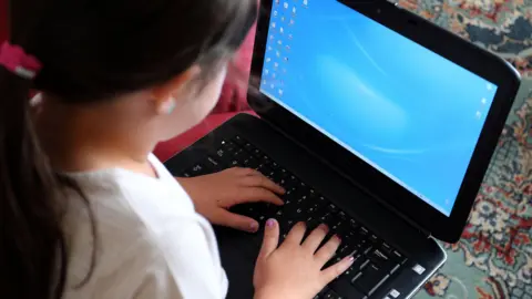 PA Media A small girl with a pony tail hairstyle and pink nail varnish uses a laptop.