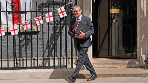 PA Media Sir Keir Starmer, in grey suit with white shirt and blue tie, is leaving 10 Downing Street, carrying folders. He is smiling and is surrounded by St George's Cross flags and bunting.