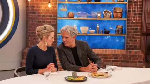 A woman with blonde hair tied up, wearing large earrings, a necklace chain, a bracelet and a dark blue long-sleeved top, with red nails is sat at a table with a man with grey hair, wearing a grey blazer and a black T-shirt. They are turning to talk to each other, with cutlery, napkins, glasses of water and two plates of food on the table in front of them. There are shelves with cooking equipment on a brick wall behind them.