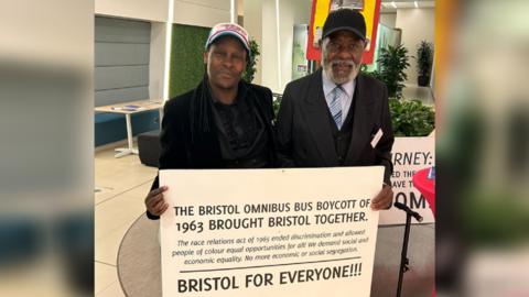 Creativist Julz Davis and Bristol Bus Boycott participant Guy Bailey holding a sign with details of the boycott which reads "Bristol for Everyone!"
