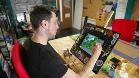 Nich Angell wearing a black t-shirt and sitting at a wooden desk in his art workshop. He is drawing a female comic book character on an iPad. 