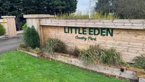 The photo shows an entry to a county park. There are wooden fences, which have the words 'Little Eden Country Park' written on them. There are numerous bushes that have been planted in front of them.