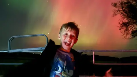 Youngster Aaron Paliczka has a big smile on his face as the aurora appears in colours of green and pink behind him.