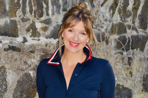 Leah Bryce A smiling woman, wearing a dark blue top, is standing with her hands behind her back in front of a stone wall. 