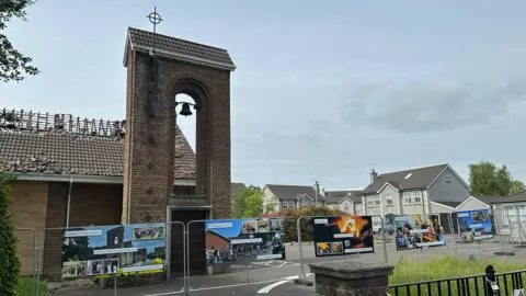 Large coloured images showing the journey of the church from the day before the fire, to the night of the fire, and the aftermath are hanging on a metal railing around the burned down church building. You can see the roof of the church building has fallen in.