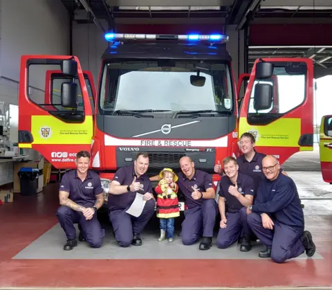 County Durham and Darlington Fire and Rescue Service Beatrix and a group of firefighters pose in front of a fire truck. The blue lights of the truck are on. They are all smiling at the camera, some have their thumbs up.