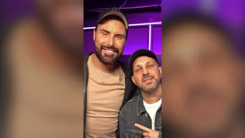 Rylan with Dynamo in the purple-lit radio studio, both wearing caps, Dynamo pointing at Rylan as they pose for a photo.