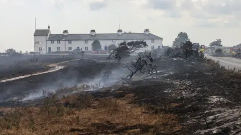PA Media A row of white cottages is in the background, and there are several emergency service vehicles parked in front of them. In the foreground, fields have been burnt due to a large fire which is still burning.