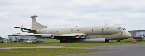 Kevin Connor A large plane beige in colour sits on grass next to area of tarmac.