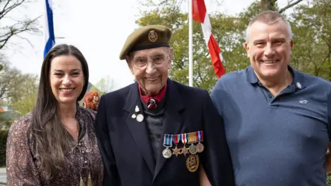 Arjan Vrieze Photography 2026 An elderly man wearing a military blazer with medals on his chest stands in the middle of a woman with long dark hair wearing a brown patterned dress and a man wearing a grey-blue shirt. They are outdoors with two flagpoles behind them and beyond that some trees in leaf.