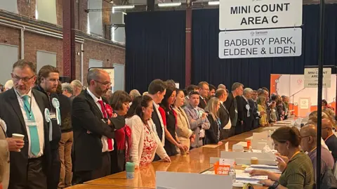 A row of trestle tables, people sitting counting votes and on the other side, politicians with rosettes standing watching.