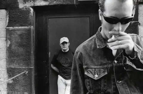 Soma Records A black and white photo of two men standing in front of a brick building. One is closer to the camera holding a cigarette in his mouth and the other is behind standing in front of a door.
