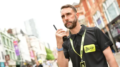 A person standing on a city street holds a handheld radio close to their mouth. The person is wearing a black polo shirt and a lanyard with a bright yellow badge displaying the text "Leeds BCRP." The background is blurred, showing urban buildings, including a tall modern tower and brick-fronted structures, creating a busy city-centre setting. The image is brightly lit and focused on the person and radio, with the surrounding street scene out of focus.