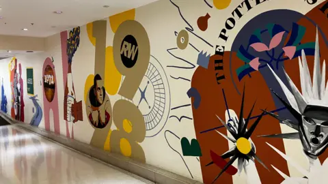 A wall that has multiple murals painted featuring different prominent years in the History of Stoke-on-Trent. There is one specifically of Robbie Williams where his face appear in the numbers of 1998 referring to the year he got his first number one single.