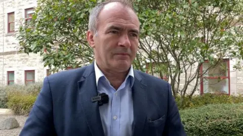 BBC Alfred Cannan, who is wearing a dark blue suit with a white shirt.
He is standing outside the Noble's Hospital building and a tree and green bushes can be seen in the background.