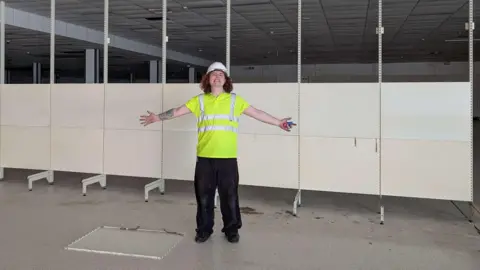The Market Gallery Theodore Godfrey-Cass stands in front of wall partitions which had been put up in the new art gallery before its planned opening. He is wearing a white hard hat and hi-vis jacket.
He has his arms spread wide and he is gurning at the camera.