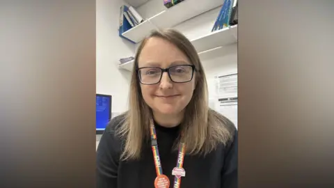 Emily Truscott A mid-shot of Emily smiling to camera. She has long brown hair and black-rimmed glasses and is wearing an NHS rainbow lanyard. She is in an office with a screen just visible in the background and A4 pages of clinical notes pinned to the back wall.