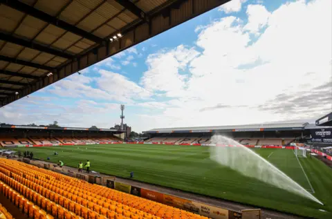 Stuart Leggett/MI News/NurPhoto via Getty Images Inside of the Port Vale football stadium. A corner angle shot showing yellow seats, the pitch, and a spray of water coming from a sprinkler.
