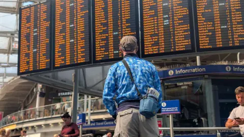 Network Rail A rail passenger seen from behind wearing a brown hat, a blue sports top and grey shorts with a blue man-bag with a newspaper sticking out of it. He stands and looks at the live information display screens in a busy railway station. There is an information desk to his right. Other passengers are in the background.