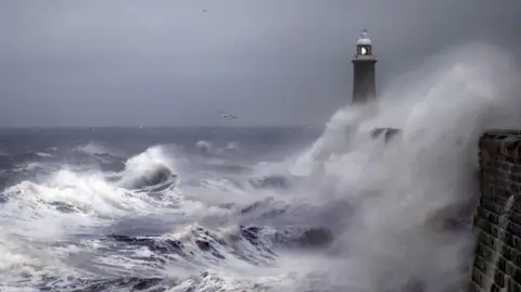 A lighthouse being splashed with large waves, there are seagulls and a cloudy darkening sky