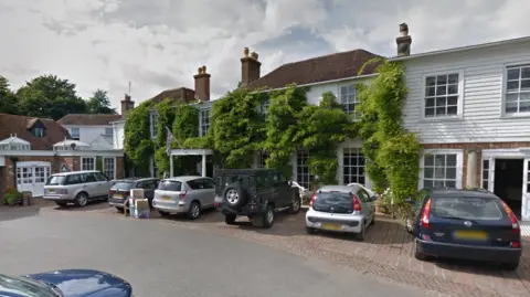 A Google Street image of the outside of the PowderMills hotel. It is an 18th century county house with white wooden cladding and red brick.