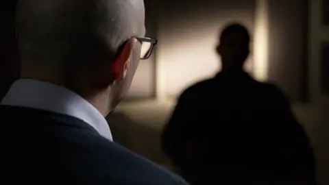 A picture of Pavan talking to Martin Jones. Pavan is blurred and sitting in darkness so you cannot identify him and Martin is sitting with his back to the camera.