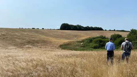 Site of the proposed forest at Hagworthingham. Two people stand with their back to the camera in the foreground in front of a small slope which is covered in yellow grass