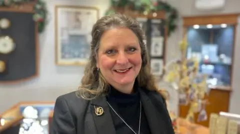 Rachel Tribbeck stands in her jewellers shop. She is surrounded by display cabinets and items for sale. She is wearing a dark suit with a brooch and necklace and is smiling to the camera.